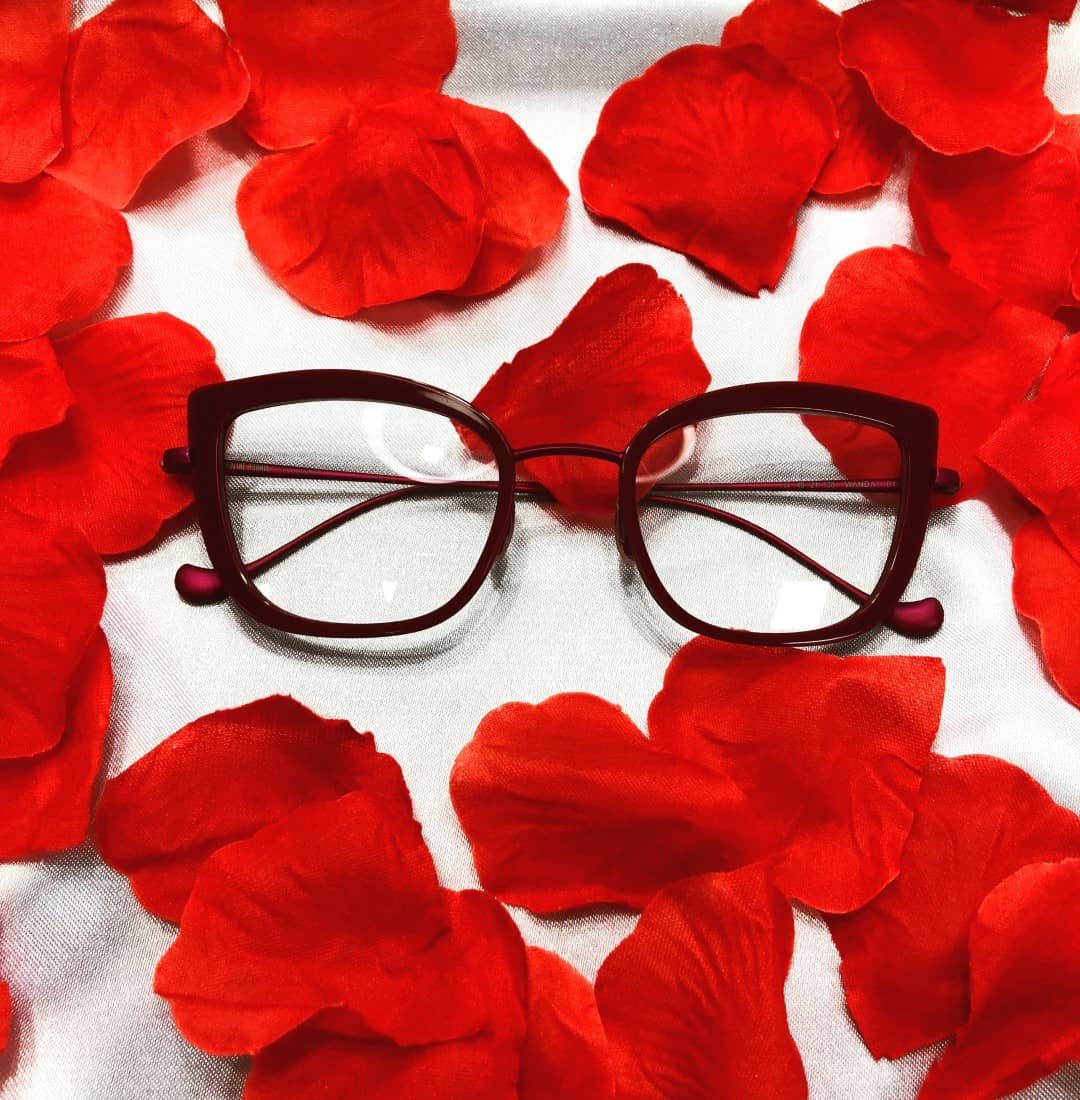 Eleganckie damskie okulary leżące na białym materiale obok czerwonych płatków kwiatów.