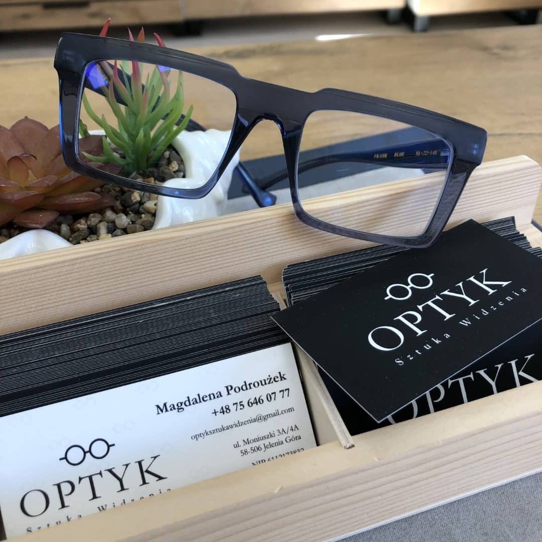 Wyjątkowe okulary na drewnianym pudełku, elegancko prezentujące na tle wizytówek salonu.