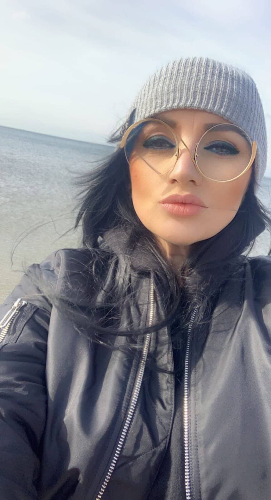 Klientka w okularach i czapce beanie na plaży, z uśmiechem na twarzy, ciesząca się słońcem i relaksującą atmosferą.